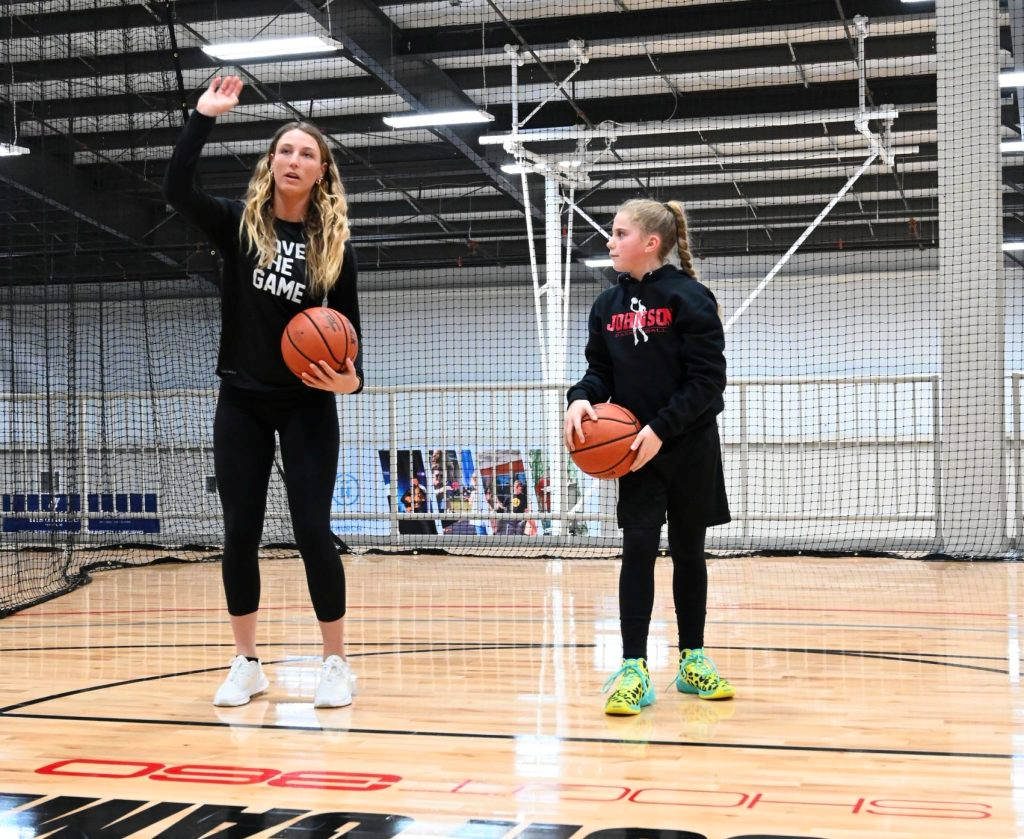 An adult and a child holding basketballs stand on an indoor court. The adult raises a hand while instructing, and the child listens attentively. Both wear athletic clothes and sneakers.