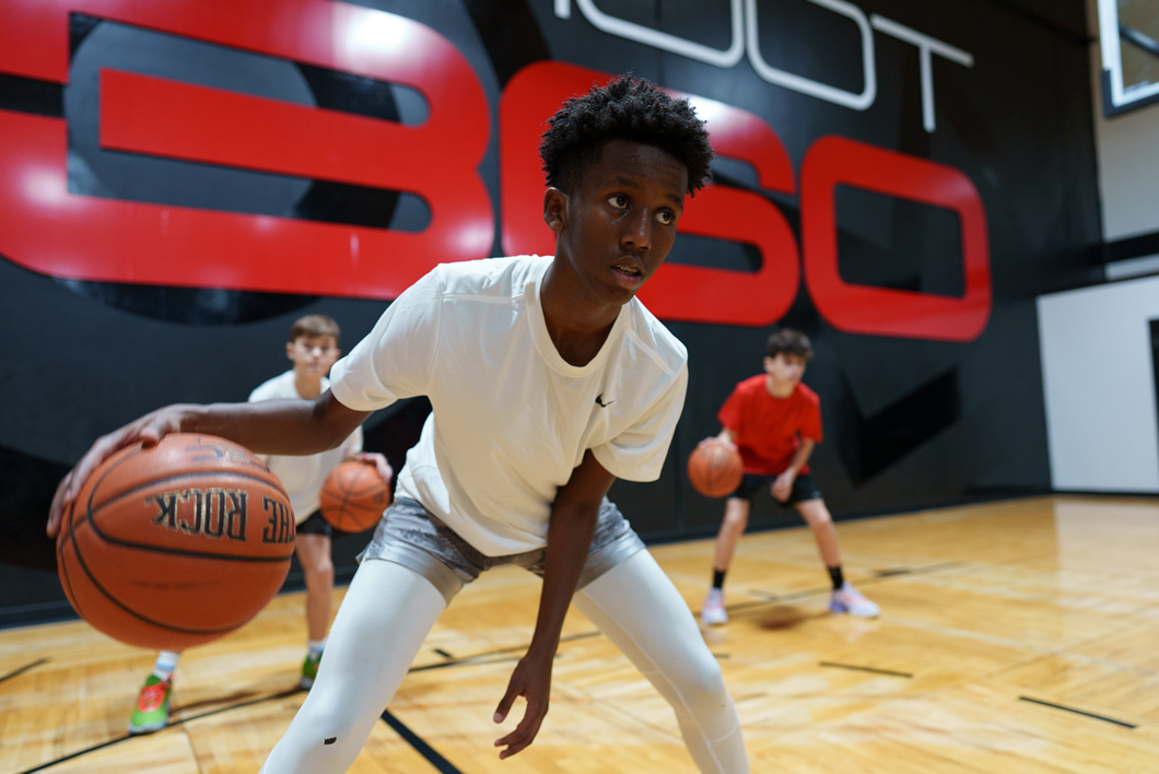 Three boys practice basketball dribbling drills in a gym. The boy in front wears a white shirt and shorts, with two others behind him in red and white shirts. The background features large red and black graphics.