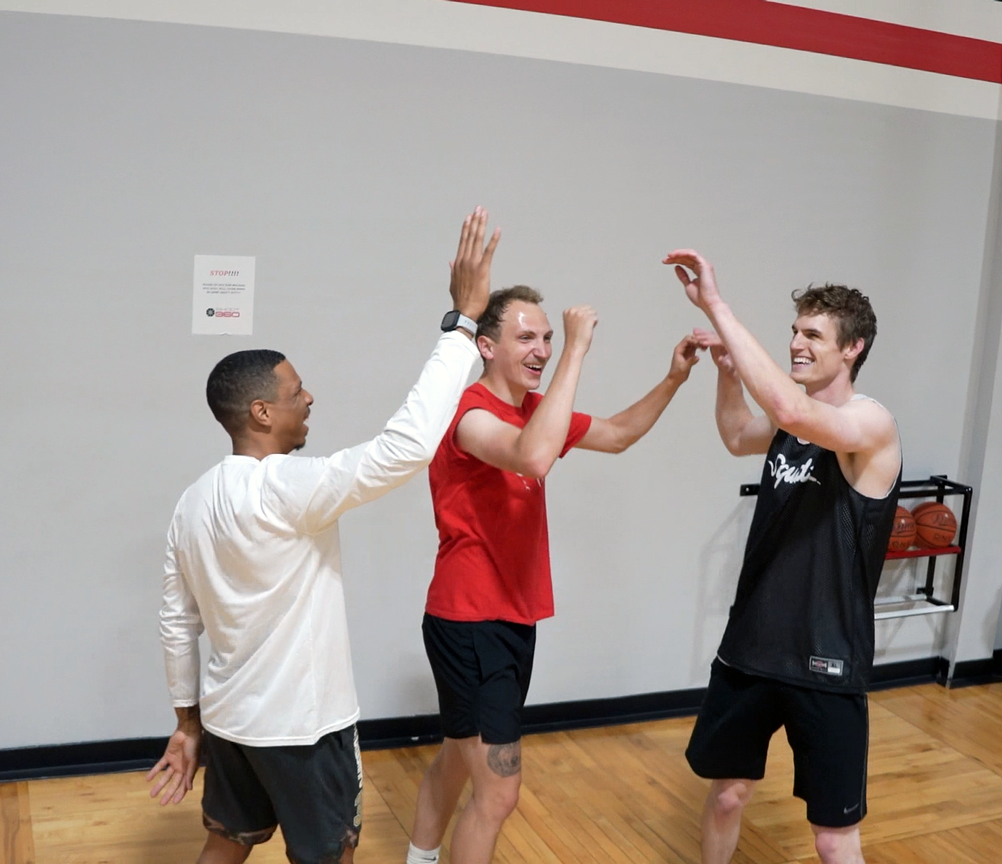 Three men in athletic wear celebrate on a basketball court, smiling and giving high fives. Basketballs and a rack are visible in the background against a white and gray wall with a red stripe.