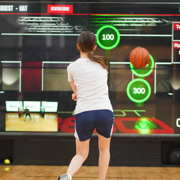 A person in sportswear shoots a basketball at a digital arcade game, aiming for targets displayed on a large interactive screen showing scores and timers.