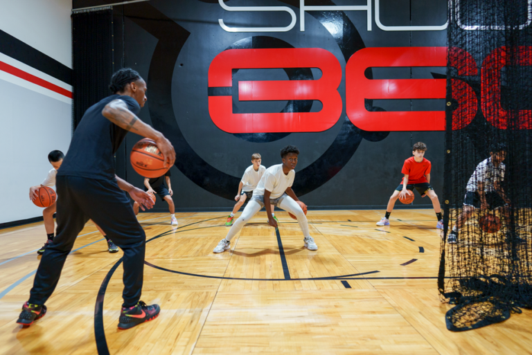 A basketball coach instructs a group of kids dribbling basketballs on an indoor court, with a large red and black wall design in the background.