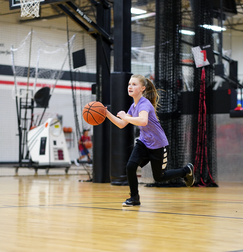 A young girl in a purple shirt and black pants dribbles a basketball on an indoor court, appearing focused and in motion. Gym equipment and a basketball hoop are visible in the background.