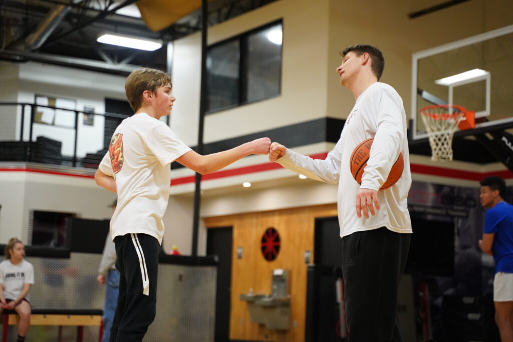 Two people in a gym fist bump, one holding a basketball. Both wear white shirts and black pants. A basketball hoop is in the background, and others sit nearby. The scene suggests teamwork or coaching.