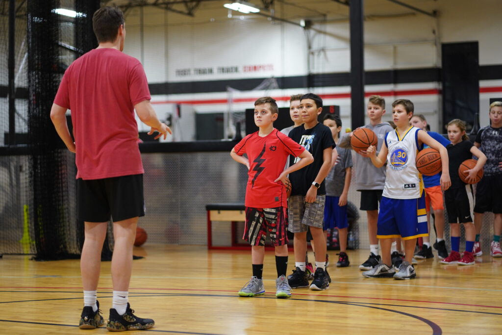 A group of boys holding basketballs lines up in a gym as a coach in a red shirt and black shorts gives instructions.