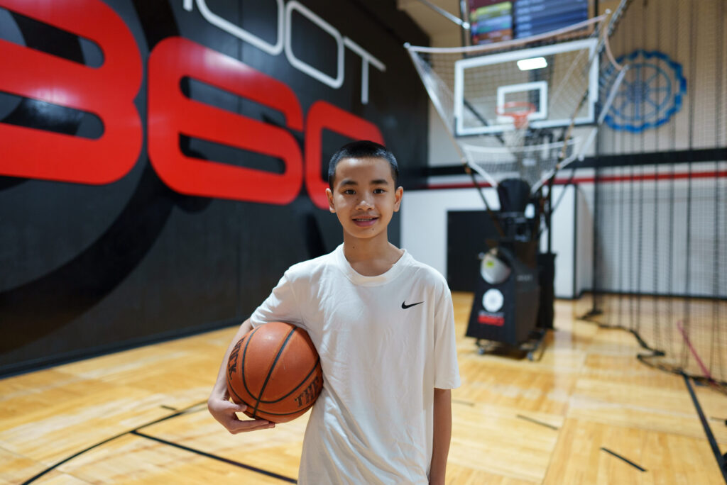 A boy in a white shirt holds a basketball on an indoor court, standing in front of a basketball shooting machine and a black and red wall.