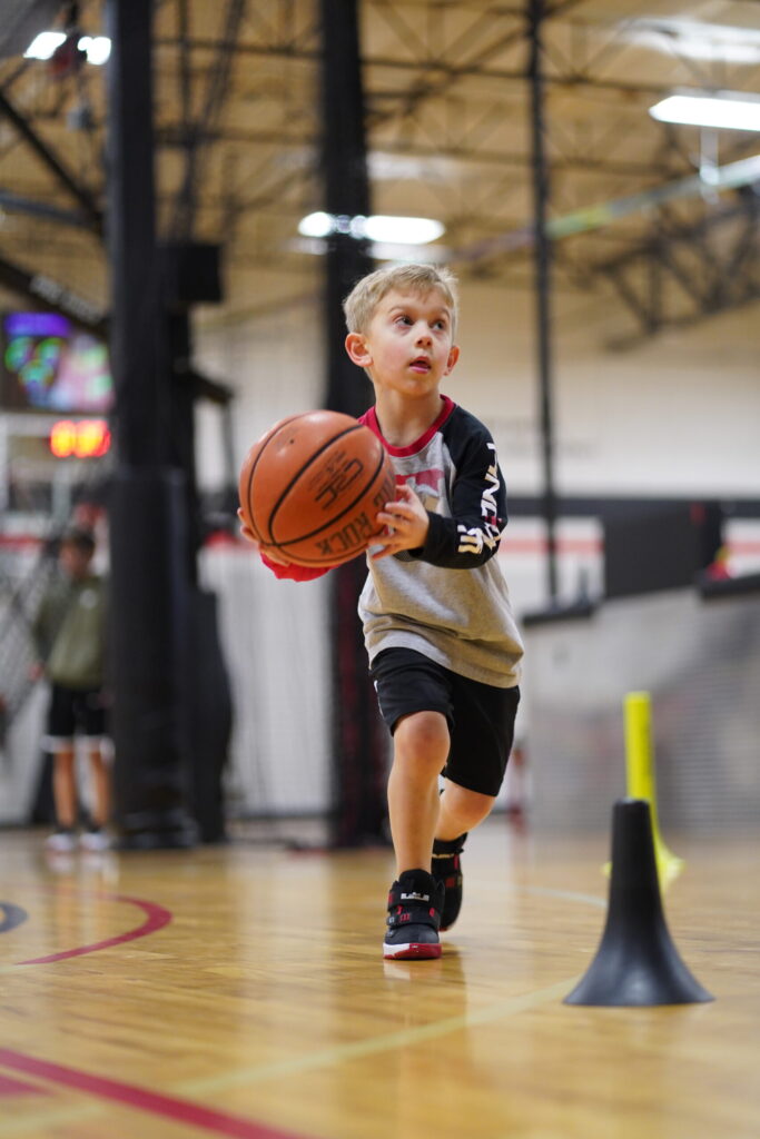 A young boy in a gray and black shirt and sneakers dribbles a basketball on an indoor court, focused on the game. Training cones are set up on the floor nearby.